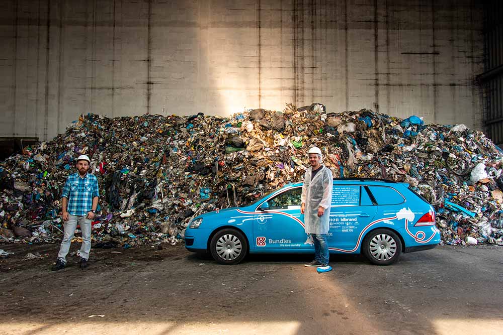Wiebe Wakker met zijn auto samen voor een berg afval, met een medewerker van een afval recycling fabriek waar Wiebe op bezoek is.